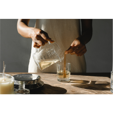 A woman pouring candle wax into a glass jar whilst holding the wick steady